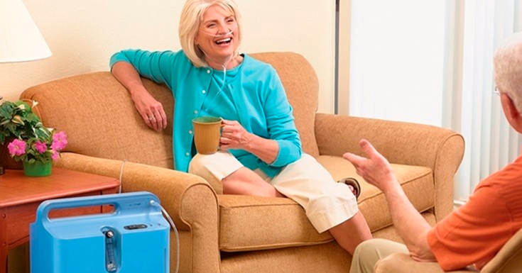 Mulher sentada na poltrona com oxigênio domiciliar, tomando café e conversando com o seu parceiro.